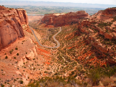 [View look down a canyon toward town. In the middle of the canyon is a swath of pavement as the road winds through it an up the canyon wall. Canyon is mostly reddish rock but also contains quite a bit of low-level vegetation.]